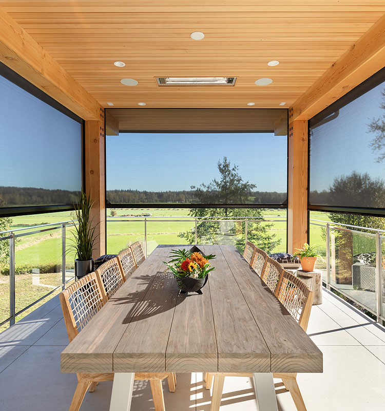 Covered patio with motorized screens and wooden dining table, overlooking green landscape through glass railings